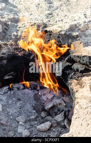 Flames emerge from a rocky crevice, contrasting with surrounding ash, dirt, and stone. Sunlight casts shadows, highlighting textures. Stock Photo