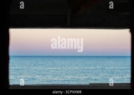 Ocean view through dark rectangular opening, featuring pink and blue pastel sky at sunset. Stock Photo