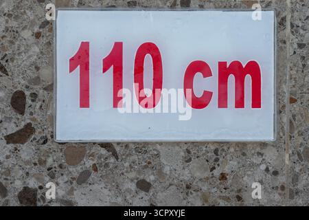 A close-up shot of a sign displaying "110 cm" in red on a white rectangular background, mounted on a speckled surface. Stock Photo