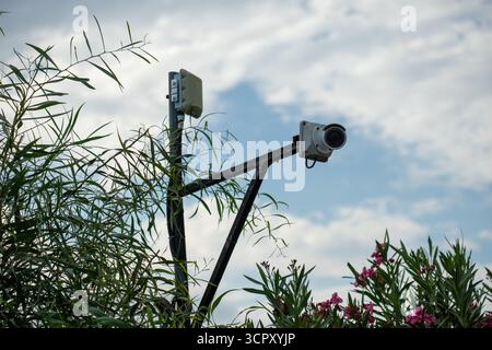 Image shows a surveillance camera mounted on a pole with a cloudy sky as a backdrop, partially obscured by foliage. Stock Photo