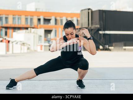 Young female doing warming-up exercises on the roof. Woman in sportswear warming up her legs. Stock Photo
