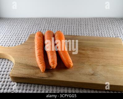 Three fresh orange carrots sit neatly on a wooden cutting board. The soft texture of the board contrasts with the vibrant color of the carrots, ideal Stock Photo