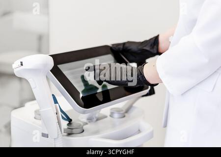 Cosmetologist in white coat and black gloves using touchscreen control panel of modern laser machine in beauty clinic, professional aesthetic equipmen Stock Photo