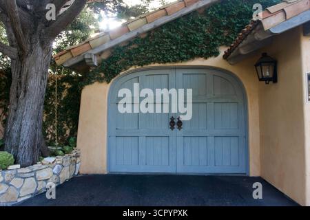 Editorial image of a rustic blue wooden garage door set into a stucco wall with ivy vines in Carmel, California. A large tree and stone planter frame Stock Photo
