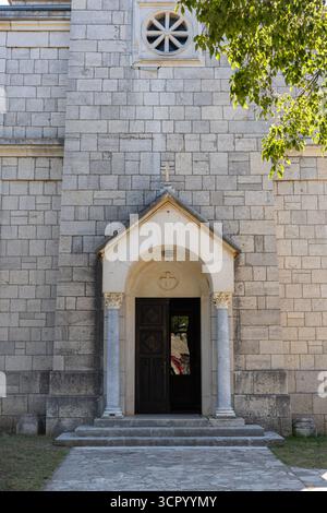 A historic stone church façade features an arched doorway, decorative columns, and a circular window above. Sunlit exterior with lush tree branches cr Stock Photo