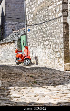 A bright red scooter rests along a sunlit, cobbled street framed by stone walls in a historic village. rustic textures, narrow lanes, and warm light e Stock Photo