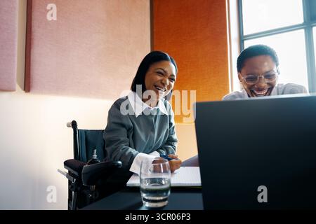 A cheerful and collaborative scene with two young women, including a wheelchair user, happily working together on a project using a laptop. Inclusivit Stock Photo