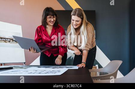 Two young women sharing ideas in a contemporary environment, reviewing design plans at a table. Stock Photo