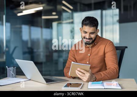 Young smiling indian businessman sitting at a desk in a modern office, concentrating on taking notes in a notebook while working with a laptop in a productive workspace Stock Photo