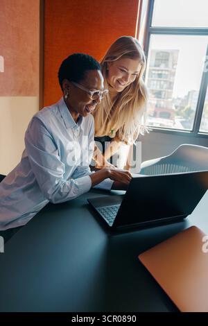 Two women of diverse backgrounds and ages collaborating at a desk in an office. The scene highlights cooperation, technology usage, and a bright work Stock Photo