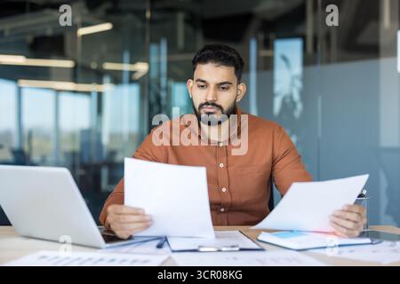 Young man analyzing financial reports and paperwork, using a laptop, and working diligently in a modern office environment, focusing on professional tasks Stock Photo