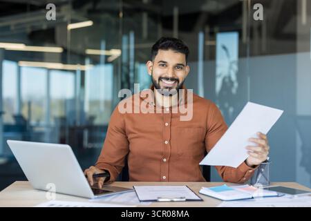 Smiling indian businessman sitting at office desk, reviewing financial documents and using a laptop, representing success, productivity, confidence, and modern business Stock Photo