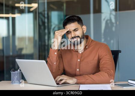 Young businessman experiencing a painful headache and severe eyestrain while working long hours on a laptop in a modern office, symbolizing stress, overwork, and burnout in professional life Stock Photo
