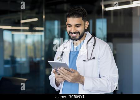 Indian doctor wearing a white coat and stethoscope, smiling while using a digital tablet in a modern hospital or clinic setting, representing medical innovation and confident healthcare Stock Photo