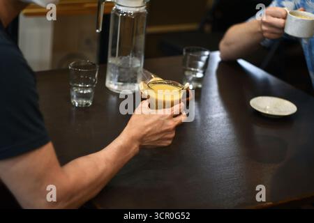Two people enjoying coffee together at a wooden cafe table. One holding an iced coffee glass, another with a hot drink, surrounded by water glasses. Stock Photo