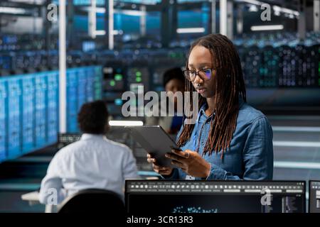 Data center admin using tablet to monitor system performance, identifying bottlenecks. Woman in server room using device to troubleshoot errors, implementing optimizations Stock Photo