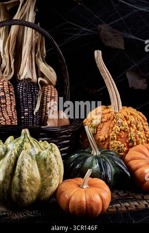 Low‑key autumn still life with a warty gourd, mini pumpkins and ornamental corn in a wicker basket, dried leaves and cobwebs against a dark background Stock Photo