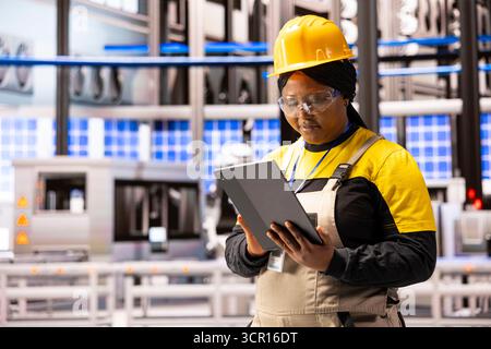 African american woman managing industrial operations with tablet, focusing on efficiency and quality control. Black female employee using digital device to test machines in smart factory. Stock Photo