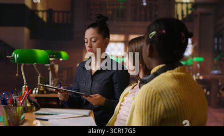 Female mentor providing instruction to diverse pupils during private lessons at the library, assisting with homework and school projects in the study area. Tutoring session for guidance. Camera B. Stock Photo