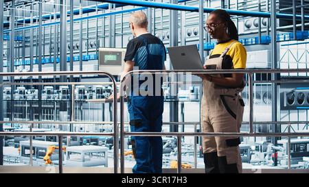 Smart factory engineer oversees operation of robotic arms and conveyor systems on facility floor. Industry 4.0 plant technician monitors system performance and troubleshoots issues, camera A Stock Photo
