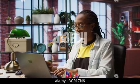 Teleworker analyzing data on laptop at desk and looking over paperwork, compiling analytical reports. Remote employee checking documents, using notebook to upload company information online, camera A Stock Photo