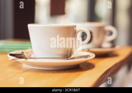 Two white coffee cups rest on small plates on a wooden table in a cozy cafe. Soft lighting creates a warm atmosphere perfect for enjoying a calm after Stock Photo