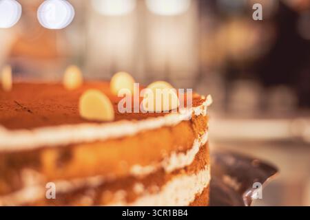 This close-up view showcases a delicious layered cake with creamy frosting and white chocolate accents, invitingly presented in a dessert shop's warm Stock Photo