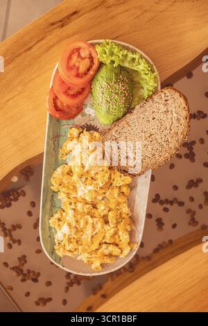 Scrambled eggs are served on a decorative plate along with fresh tomatoes, lettuce, avocado, and whole grain bread, creating a cozy breakfast scene on Stock Photo