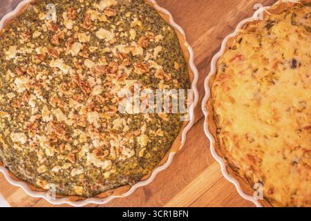 Two freshly baked quiches rest on a wooden surface, one filled with greens and topped with nuts, while the other is loaded with cheese and vegetables, Stock Photo