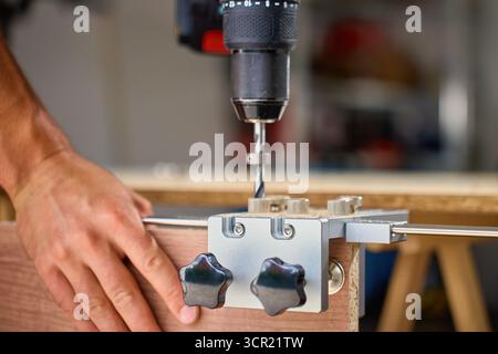 Close-up of carpenter drilling hole in wooden panel using drill and metal jig tool for precision. Man working in home workshop. Concept of woodworking Stock Photo