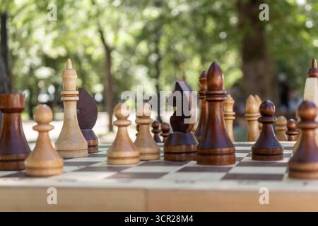 The image presents a close-up view of a chessboard set up in an outdoor setting, likely a park, during daylight hours with focus on the chess figures Stock Photo