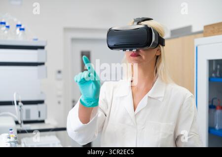 Female researcher wearing VR headset in a high tech chemical lab, performing virtual experiment simulation. Concept of augmented reality in science, digital data visualization, and innovative research technologies. Stock Photo