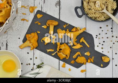 White, wooden table and yellow mushrooms on a graphite chopping board, broken egg, black pot of pasta and green pepper. Stock Photo