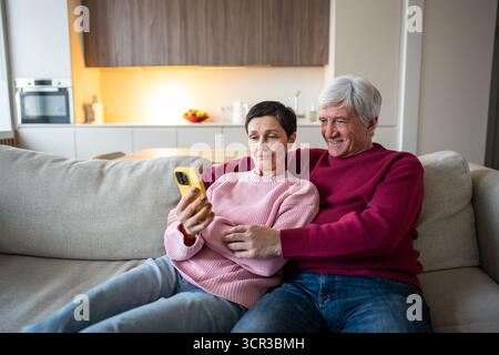 Seniors couple explore ai app on smartphone, calls their relatives via video chat, sitting on sofa.  Stock Photo