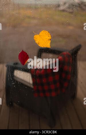 Rain falls on a window. Outside, a gray chair sits with a red plaid blanket. Two fallen leaves stick to the glass on a cool Autumn day. Stock Photo
