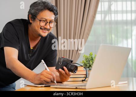 Happy adult freelance handsome photographer at home, working on laptop in home office. Selective focus. Stock Photo