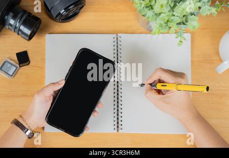 Female hands holding a smart phone and blank notebook with camera and memory cards on wooden background, flat lay. Stock Photo