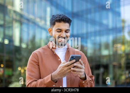 Young man smiling while browsing and texting on a smartphone outdoors in a modern urban street, enjoying social apps, connectivity and a cheerful, casual city lifestyle Stock Photo