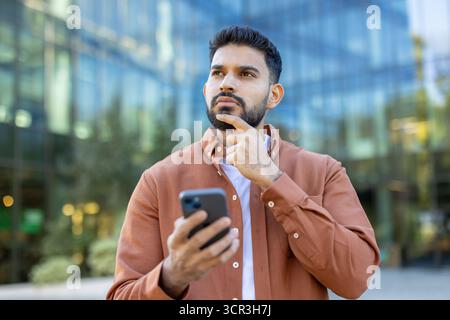 Thoughtful young indian businessman standing outdoors in a city, holding his smartphone and gazing upward in focused contemplation, planning strategy and future success Stock Photo