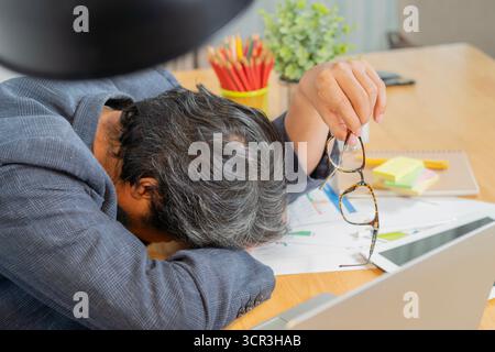 Lazy adult man sleeping in work time, male officer feeling tired and dizziness, he holding a eyeglasses while rest face on work table, overwork and ha Stock Photo