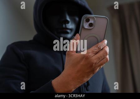 An anonymous masked hacker is using a smartphone to penetrate credit card financial information. Hacking and malware concept. Stock Photo