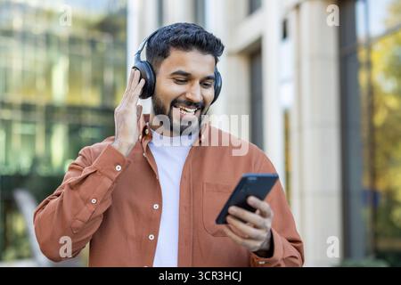 Smiling young man enjoying audio content on his smartphone, wearing wireless headphones while walking casually in an urban environment with modern buildings Stock Photo