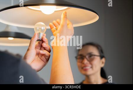 Happy loving couple changing light bulb or repairs lamp in their apartment. Family doing DIY renovations at home.  Selective focus. Stock Photo