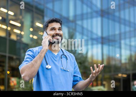 Young male doctor smiling during a phone call, wearing scrubs and a stethoscope while standing outdoors with a modern hospital or office building in the background Stock Photo