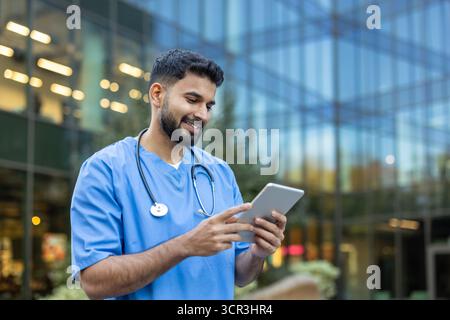Young male doctor smiling while looking at and using a digital tablet, wearing medical scrubs and a stethoscope in front of a modern hospital building Stock Photo