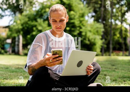 Freelance young woman multitasking with computer and smartphone in park Stock Photo