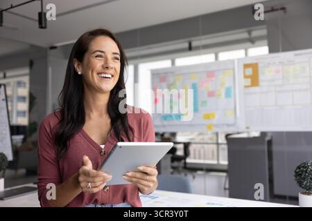 Happy office employee holding tablet, looking away with joyful expression Stock Photo