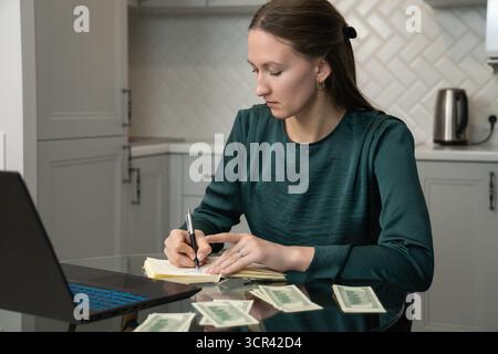 Focused woman writing notes in notebook, managing home budget, calculating money and making financial plan while sitting at kitchen table with laptop Stock Photo