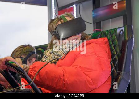 Cheerful woman with her son driving a virtual simulator in an amusement park. Blonde woman wearing a mask on a virtual simulator Stock Photo