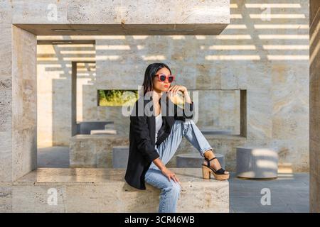 Stylish woman in sunglasses poses thoughtfully on a stone structure in a sunlit setting. Spain Stock Photo